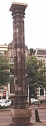 A tall, ornate column standing alone, surrounded by trees and buildings.