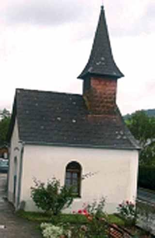 A small chapel with a pointed roof and a single window, surrounded by plants.