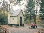 A small chapel in a wooded area with two people sitting on a bench nearby.
