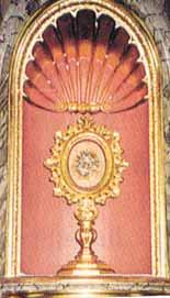 A golden monstrance displaying the Eucharistic host, set against a decorative background.