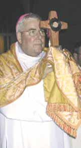 A priest holding a cross with a circular center, dressed in ceremonial robes.