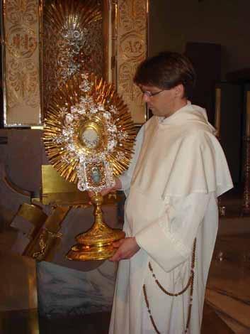 A man in a white robe holding a golden monstrance with a religious relic inside.