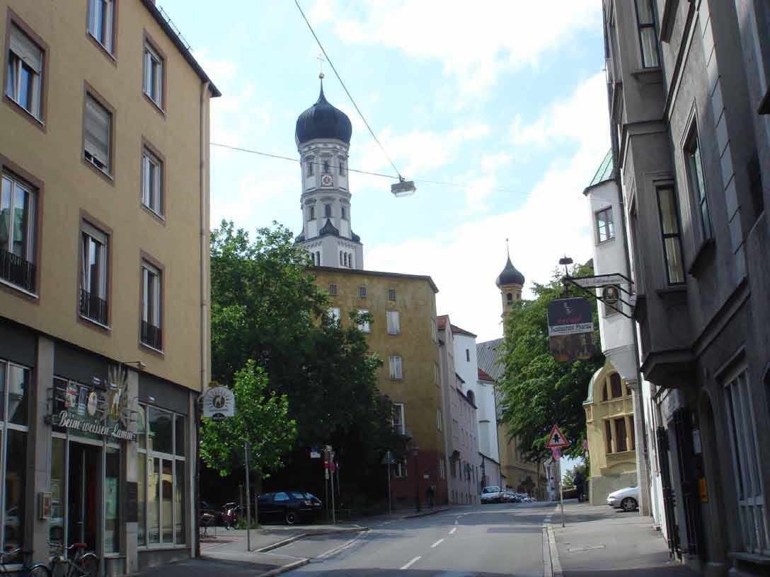 A street view in Augsburg showing buildings and church towers under a blue sky.