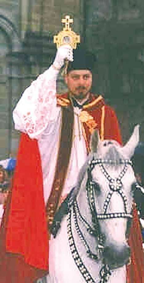 A man in ceremonial attire holds a relic while riding a decorated white horse.