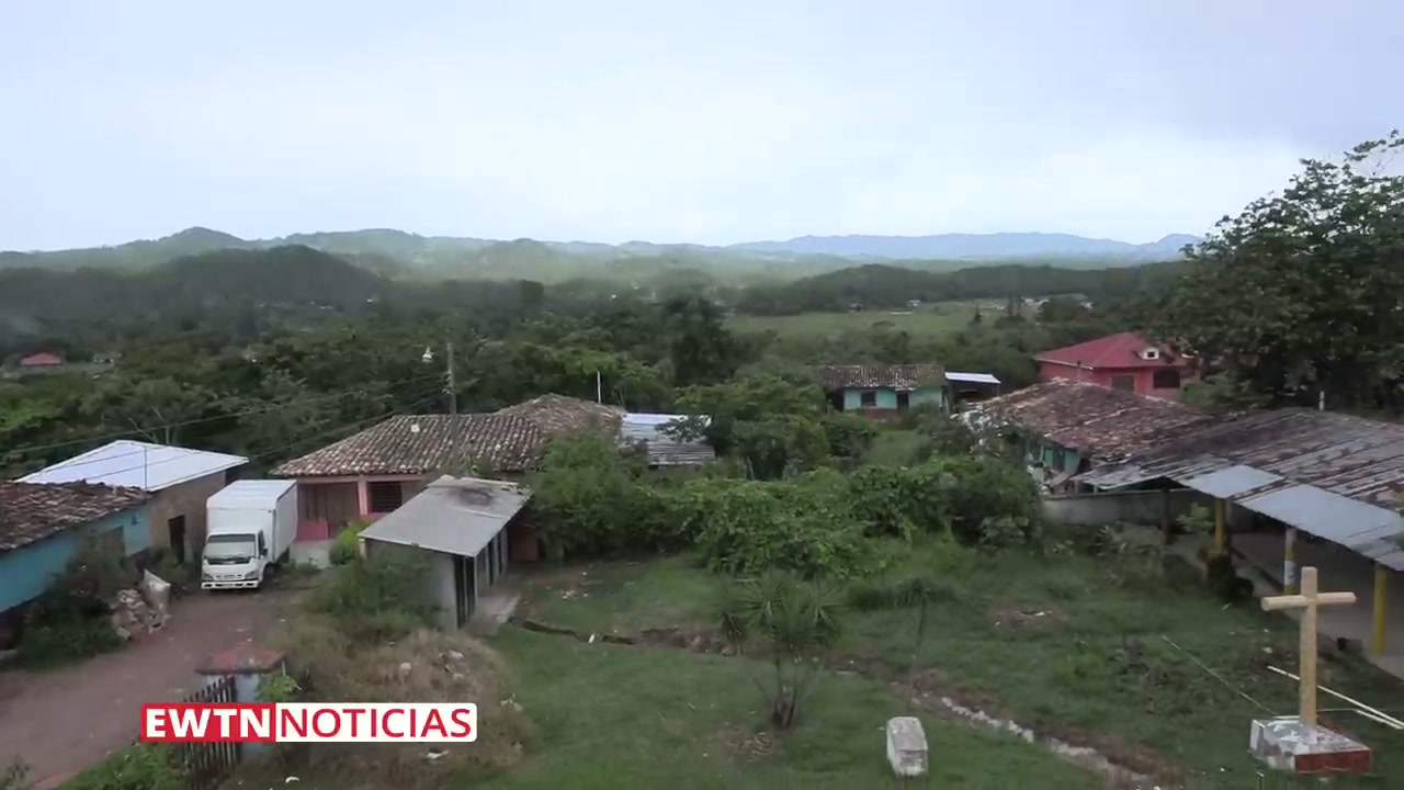A panoramic view of the rural landscape surrounding San Juan in the department of Intibucá, Honduras. Scattered houses with tile roofs sit among green hills and misty mountains, with a wooden cross visible in the foreground.