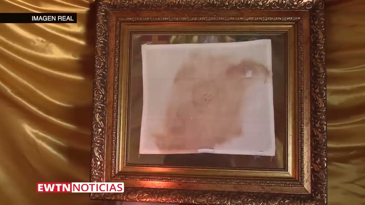 The actual blood-stained corporal, labeled 'IMAGEN REAL' (real image) in the EWTN broadcast, displayed in an ornate gold frame against gold satin. The brown-red blood stains and a small embroidered cross are visible on the white linen cloth.