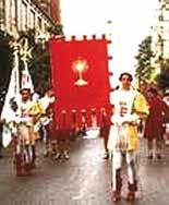 A procession with people in historical costumes carrying a large red banner featuring a chalice.