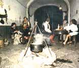 A group of people sitting around a table in a room with a pot hanging over a fire.