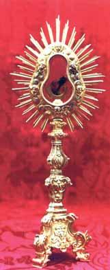 An ornate monstrance with a sunburst design, displaying a relic against a red background.
