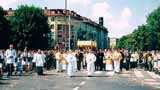 A religious procession with clergy and followers walking down a street, surrounded by buildings and trees.