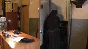 A nun in a dark habit stands near a door in a room with wooden furniture.