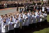 A group of altar servers in white robes waving at a large crowd during a ceremony.