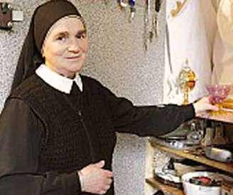 Sister Julia Dubowska stands beside a shelf, holding a vessel with a serious expression.